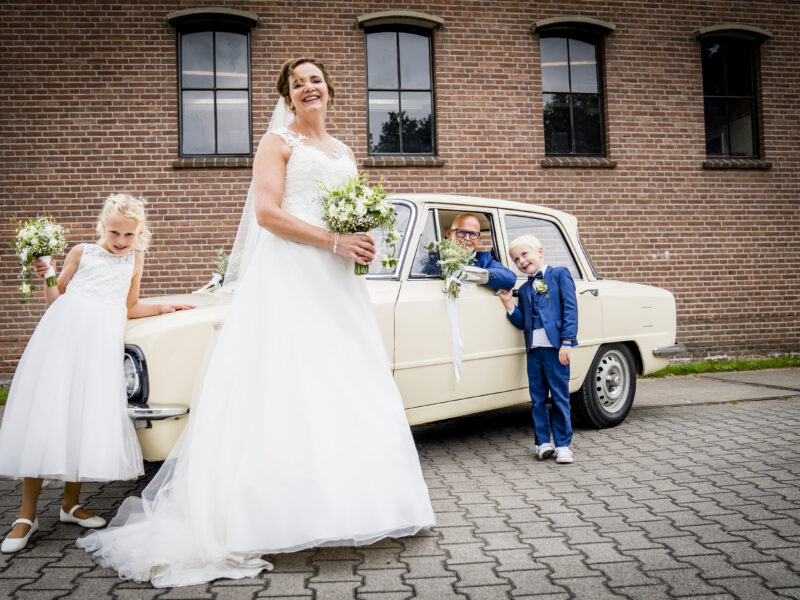 Bruidegom in auto, bruid, bruidsjonker en -meisje ervoor. Fotograaf Karin Keesmaat van Kijk-Kunst fotografie
